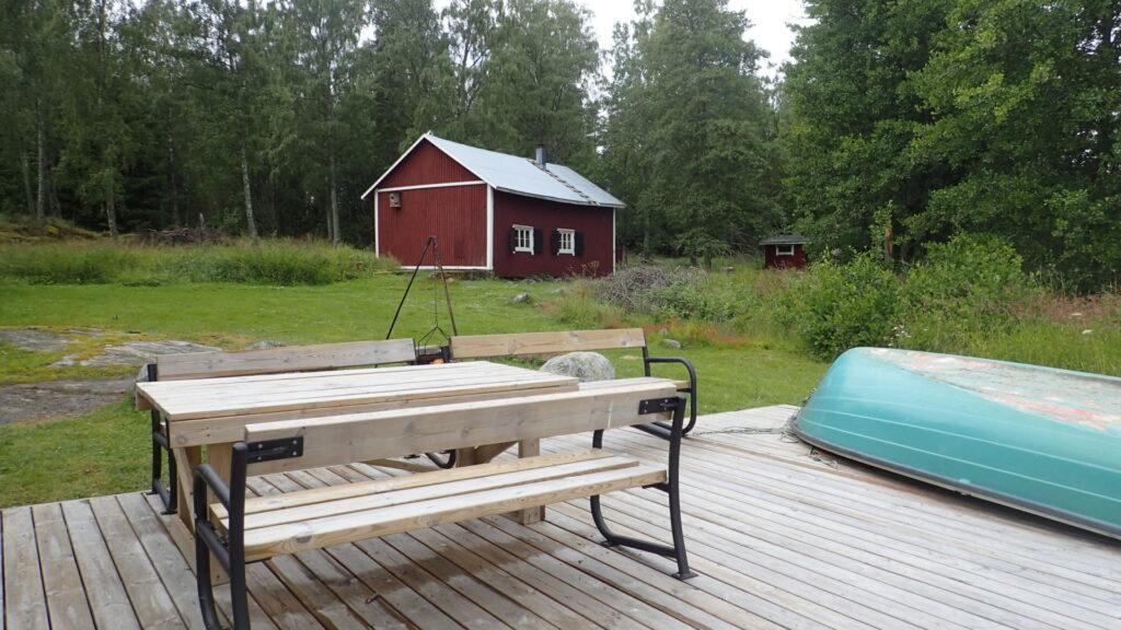 Punainen rakennus, pöytä ja istuimia sekä vene Vesiluomassa