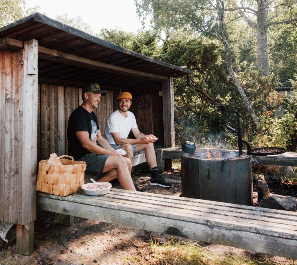 Two people sitting in a traditional Finnish lean-to shelter.