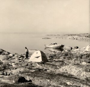 An old photograph showing a tent on a rock and a man standing next to the tent. In the background, there is the sea and a boat.